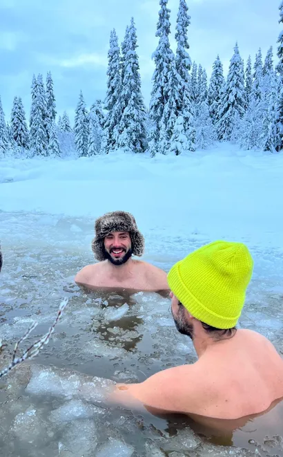 Two shirtless men in an icy geothermal pool surrounded by snow-covered evergreen trees