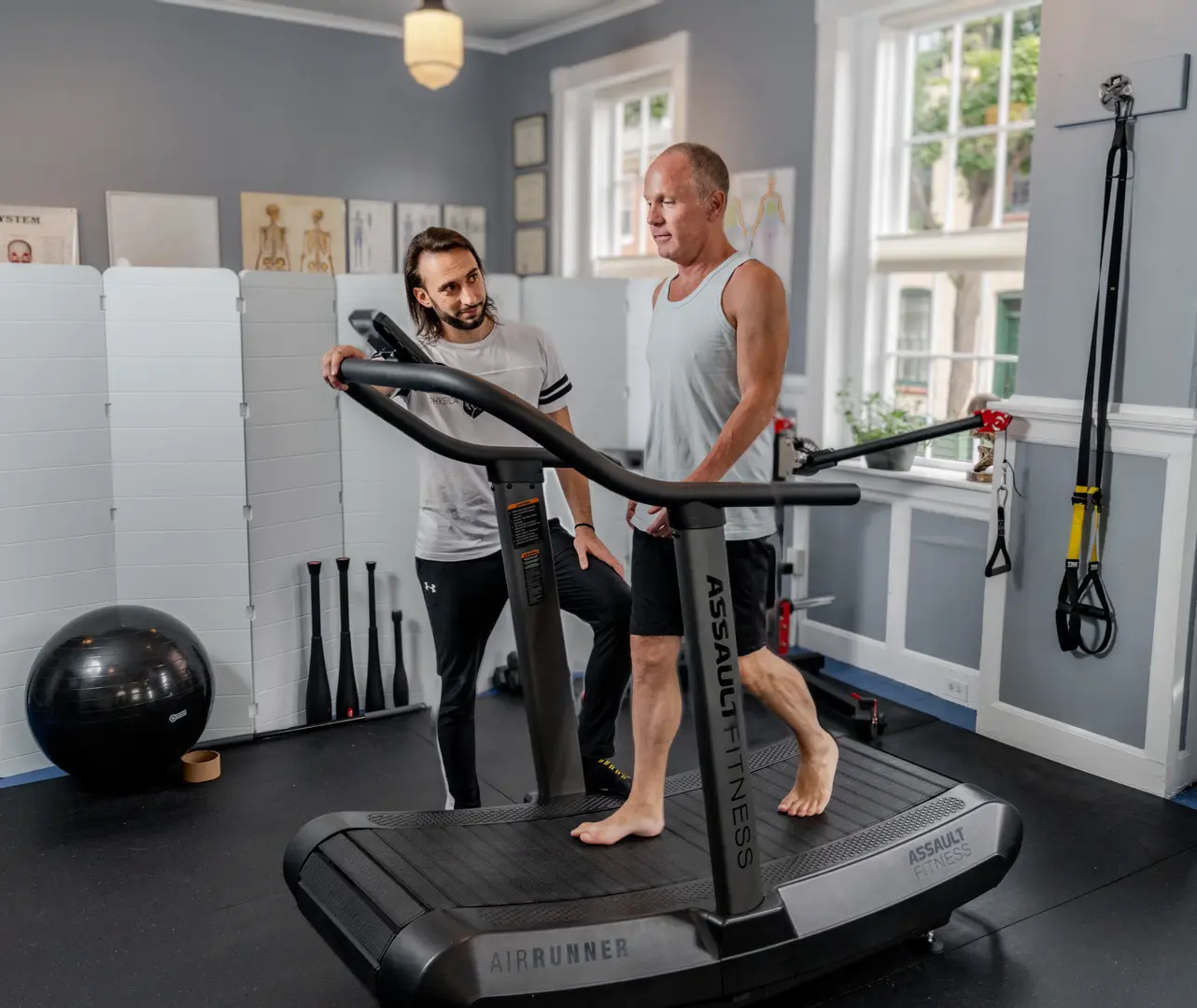 A practitioner instructs a client on proper form while using an exercise bike in a bright, modern therapy room.