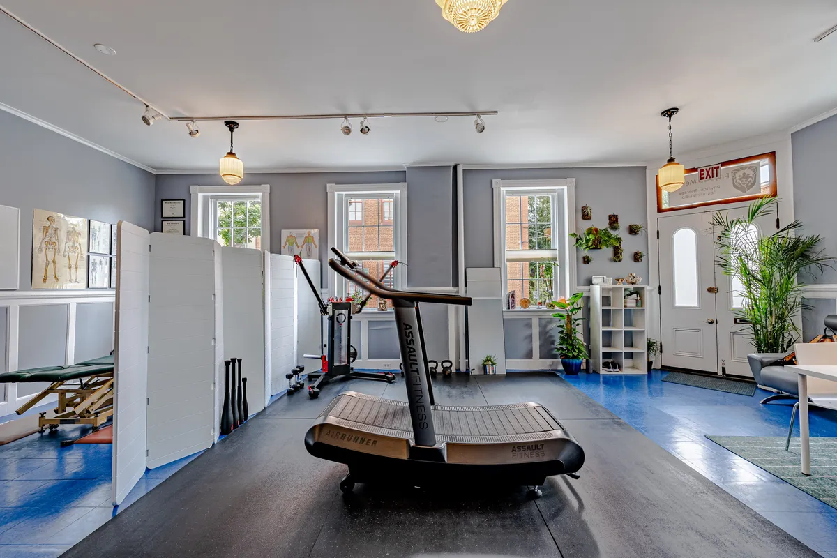 A spacious, modern therapy clinic interior featuring exercise equipment, anatomical charts, natural lighting, and professional design elements.