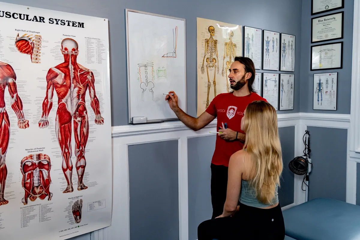 A male practitioner in red branded shirt explains anatomical concepts to a female client using wall-mounted anatomical charts and diagrams.