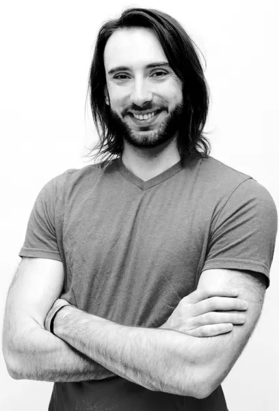 Black and white portrait of a man with long dark hair and beard wearing a gray v-neck shirt, arms crossed
